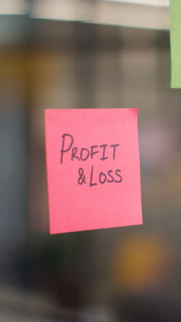 Vertical-Video-Close-Up-Of-Woman-Putting-Sticky-Note-With-Profit-And-Loss-Written-On-It-Onto-Transparent-Screen-In-Office-